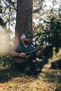 Nate Michaels playing guitar sitting against a tree.