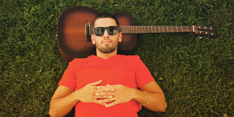 Kevin Ludwig wearing sunglasses laying in the grass with the guitar behind his head as a headrest.