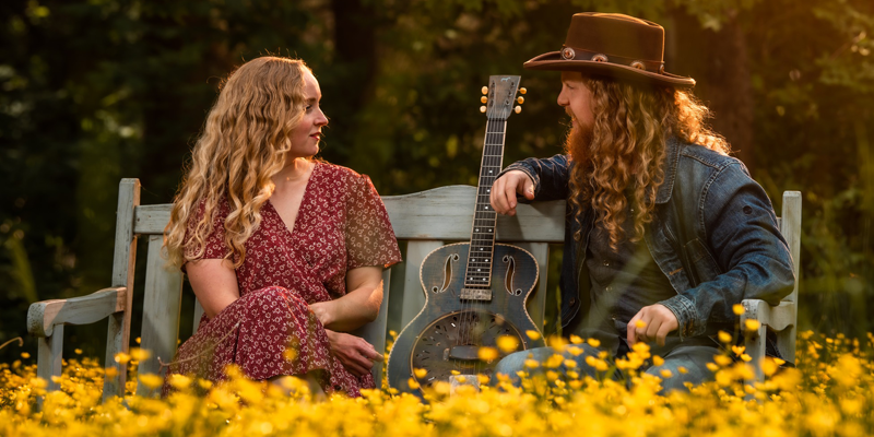 Kody and Herren sitting on a bench in a field of yellow wild flowers with a guitar.